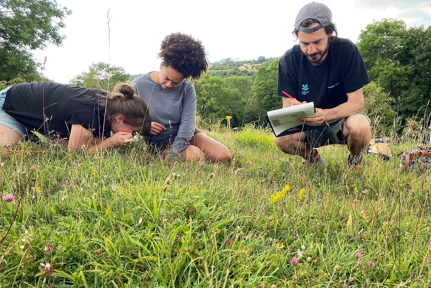 A Food Partnership staff sit in the grass to get a closer look at the wild flowers with a magnifying loupe, noting down what they find.